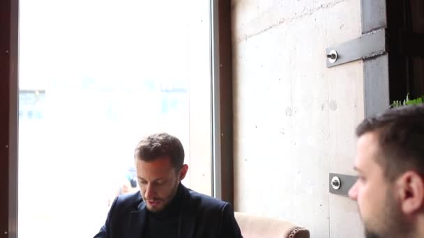 Dos hombres de negocios sentados en la mesa de la cafetería, usando el ordenador portátil, tomando té y hablando — Vídeos de Stock