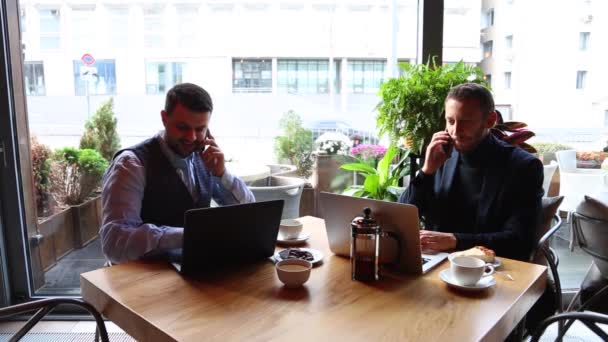 Dos hombres de negocios sentados en la mesa de la cafetería, usando el ordenador portátil, y hablando por teléfono inteligente — Vídeos de Stock