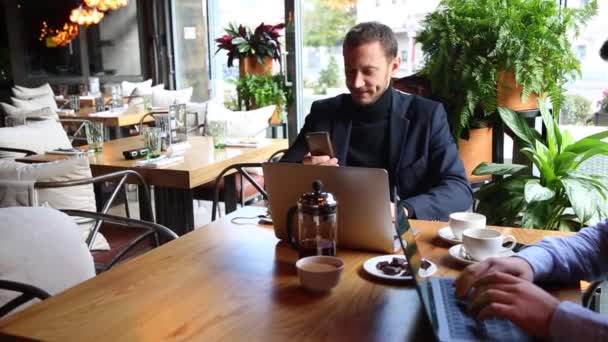 Dos hombres de negocios sentados en la mesa de la cafetería, usando el ordenador portátil, tomando té y hablando — Vídeos de Stock