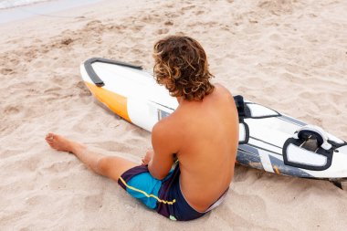 Yakışıklı adam dalga deniz okyanus sahilinde spot sörf yapmak için beyaz boş sörf tahtası beklemek ile sahilde oturuyor. Spor, fitness, özgürlük, mutluluk, yeni modern yaşam, hipster kavramı.