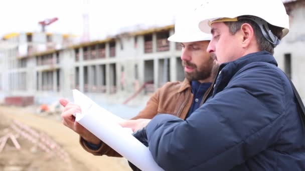 Dois engenheiros falam sobre o projeto para o edifício de construção. Eles olham para ambos os projetos de papel — Vídeo de Stock