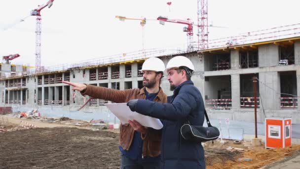 Dois engenheiros falam sobre o projeto para o edifício de construção. Eles olham para ambos os projetos de papel — Vídeo de Stock