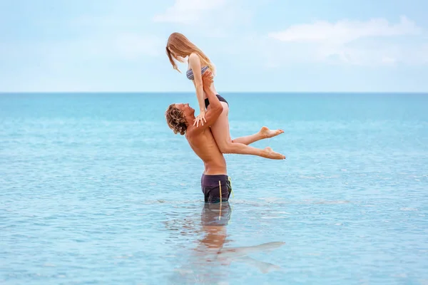 La mujer salta hacia el hombre en sus brazos, de pie en el mar. Ambos están en traje de baño. Sonriente joven pareja juguetona enamorada divirtiéndose en la playa de arena . —  Fotos de Stock