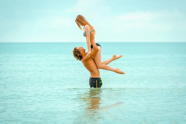 Woman jumps to the man in his arms, standing in the sea. Both are in the swimsuits. Smiling playful young couple in love having fun at sandy beach.