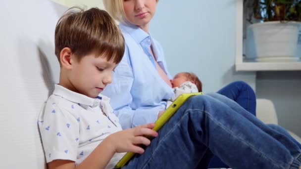 Trzyosobowej rodziny siedzą na kanapie i korzysta z urządzeń cyfrowych. Chłopiec przy użyciu tabletu i przeglądanie informacji na smartfonie podczas karmienia piersią dziecka matka — Wideo stockowe