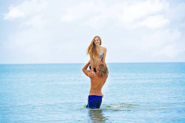 Woman jumps to the man in his arms, standing in the sea. Both are in the swimsuits. Smiling playful young couple in love having fun at sandy beach.