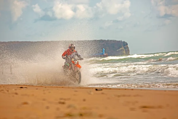 Motociclista in tuta protettiva cavalca una moto sul mare, schizzi volano da sotto le ruote . — Foto Stock