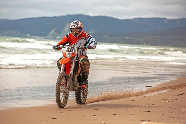 Motocyklista v ochranný oblek a jede na motorce na moře, šplouchání s poklopcem na písku pod kola. — Stock fotografie