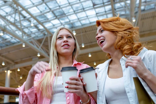Дві красиві усміхнені жінки з одноразовими чашками кави розмовляють у торговому центрі. Розмитий торговий центр як фон. Концепції способу життя та дружби . — стокове фото