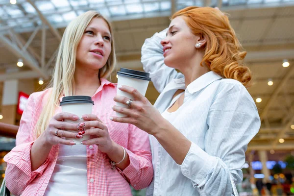 Dwie piękne uśmiechnięte kobiety z jednorazowymi filiżanek do kawy mówią w centrum handlowym. Rozmycie centrum jako tło. Koncepcje stylu życia i przyjaźni. — Zdjęcie stockowe