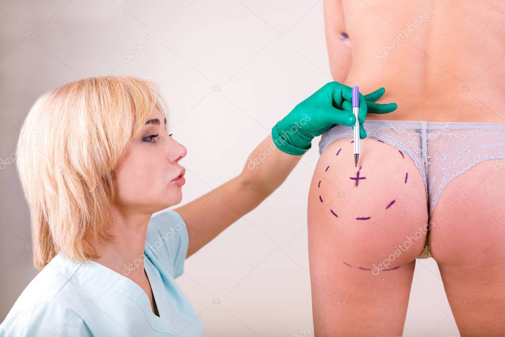 Surgeon drawing marks on female body before plastic operation, white background. Beautician touch and draw correction lines on womans buttocks.