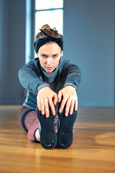 Ung smuk kvinde i sportstøj gør strækker mens du sidder på gulvet foran vinduet på gym - Stock-foto