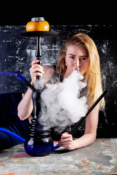 Uma mulher jovem e bonita fuma um cachimbo de água no café. Produz fumo da boca dele. O prazer de fumar . — Fotografia de Stock