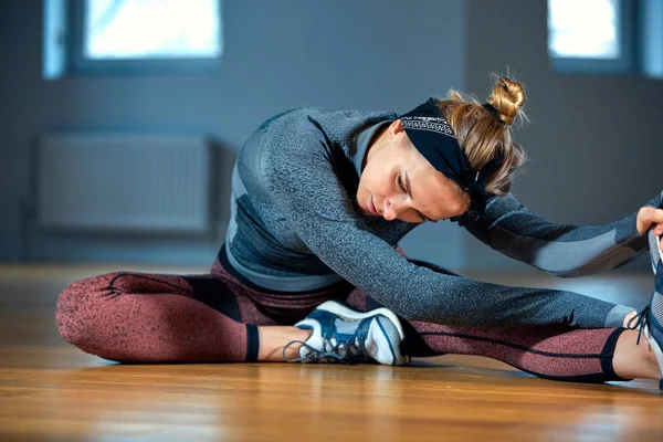 Jeune belle femme en vêtements de sport faire des étirements tout en étant assis sur le sol devant la fenêtre à la salle de gym — Photo