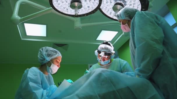 Χειρούργοι φορούν προστατευτικό ρουχισμό εκτέλεση χειρουργικής χρησιμοποιώντας αποστειρωμένο εξοπλισμό. Ιατρική ομάδα εκτελεί χειρουργική επέμβαση σε φωτεινό σύγχρονο λειτουργικό δωμάτιο — Αρχείο Βίντεο