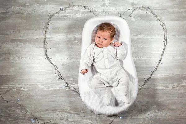 Neugeborenes schläft in einem speziellen orthopädischen Matratzen-Babykokon, auf einem Holzboden und einem Herz aus Girlanden. Ruhiger und gesunder Schlaf bei Neugeborenen. — Stockfoto