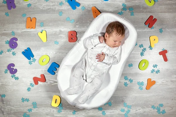 Pasgeboren baby slaapt in een speciale orthopedische matras Baby cocoon, op een houten vloer veelkleurige brieven rond. Rustig en gezond slapen bij pasgeborenen. — Stockfoto
