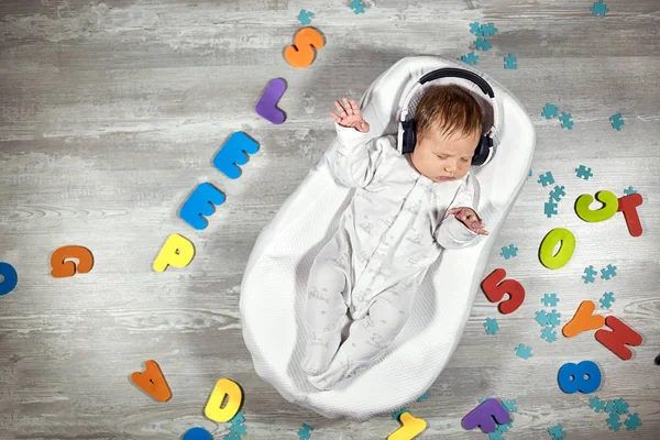 Neugeborenes schläft in Kopfhörern in einem speziellen Matratzen-Babykokon, auf einem Holzboden herumstehen bunte Buchstaben. Ruhiger und gesunder Schlaf bei Neugeborenen — Stockfoto