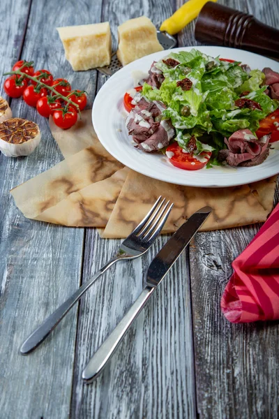 Листя салату з нарізаною смаженою яловичиною та сушеними вишневими помідорами на дерев'яному фоні — стокове фото