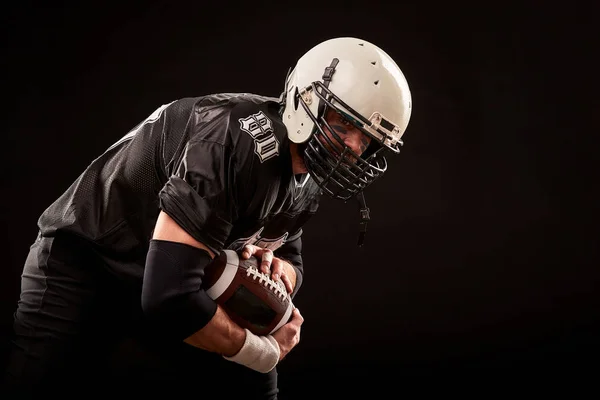 Jugador de fútbol americano en uniforme oscuro con la pelota se prepara para atacar sobre un fondo negro . — Foto de Stock