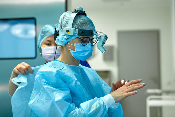 Хирурги носят стерильную одежду перед операцией. Команда хирургов готовится к операции, устанавливает дополнительный свет на голову
.