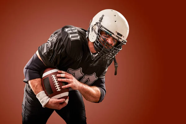 American football player in dark uniform with the ball is preparing to attack on a brown background.