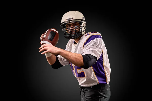 Jugador de fútbol americano en uniforme con la pelota se está preparando para hacer un pase. Concepto de fútbol americano, fondo negro — Foto de Stock