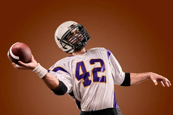 Jugador de fútbol americano en uniforme con la pelota se está preparando para hacer un pase. concepto de fútbol americano, fondo marrón — Foto de Stock