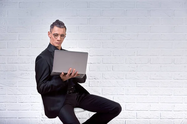 Красивий бізнесмен в окулярах і костюм тримає ноутбук в руках і щось пише. Вид збоку. На білому цегляному фоні — стокове фото