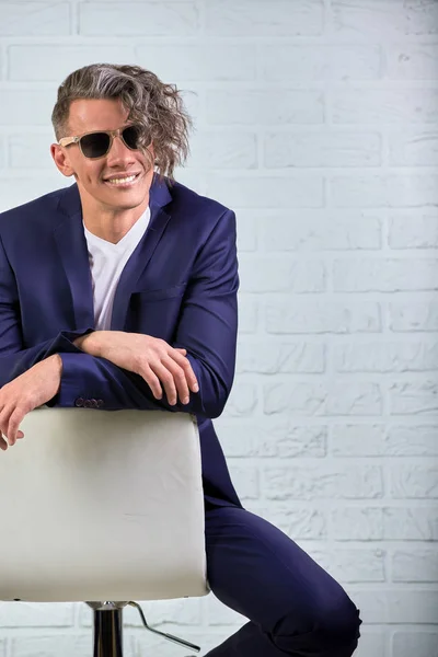Homem de negócios elegante com cabelos longos encaracolados em óculos de sol sentados na cadeira no fundo branco — Fotografia de Stock