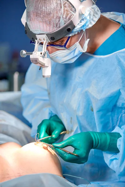 Cirurgião que realiza cirurgia de aumento mamário no centro cirúrgico do hospital. Cirurgião em máscara vestindo lupas durante a aquisição médica . — Fotografia de Stock