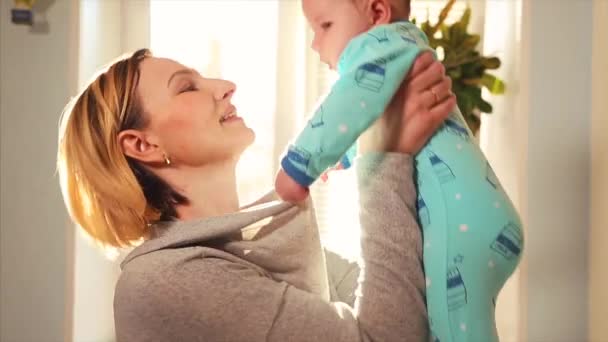 Mother plays with baby in her arms and hugs against window in su ny day — Stock Video