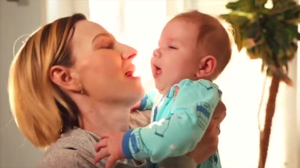 Η μητέρα παίζει με το μωρό στην αγκαλιά της και αγκαλιές κατά παράθυρο su ny ημέρα — Αρχείο Βίντεο