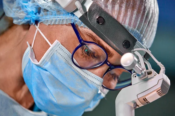 Foto do cirurgião operado na sala de cirurgia. Cirurgião em máscara e óculos com farol montado. Retrato fechado — Fotografia de Stock