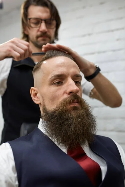 Brutal guy in modern Barber Shop. Hairdresser makes hairstyle a man with a long beard. Master hairdresser does hairstyle by scissors and comb