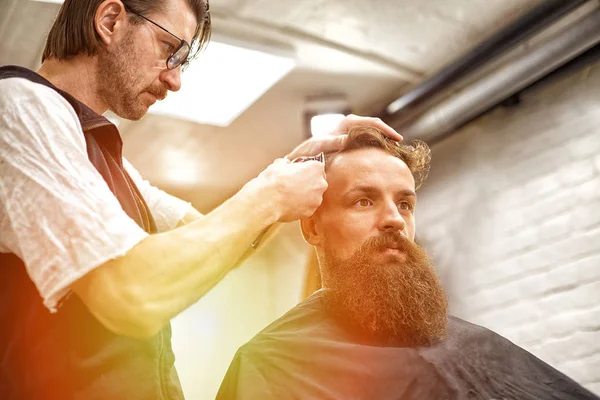 Жестокий парень в современной парикмахерской. Парикмахер делает прическу мужчине с длинной бородой. Мастер парикмахерская делает прическу ножницами и расческой — стоковое фото