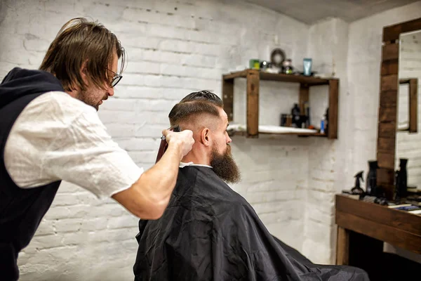 Brutaler Typ im modernen Friseurladen. Friseur frisiert einen Mann mit langem Bart. Friseurmeister frisiert mit Haarschneidemaschine — Stockfoto
