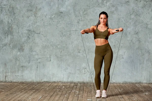 Modelo de aptidão sorridente posando no ginásio de comprimento completo contra uma parede cinza com jumpers em suas mãos, conceito Fitness — Fotografia de Stock