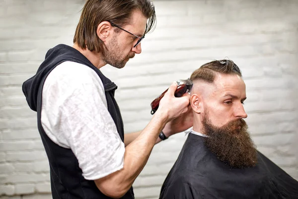 Stylish barber cuts a brutal man with a thick beard.