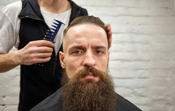 Master cuts hair and beard of men in the barbershop, hairdresser makes hairstyle for a young man