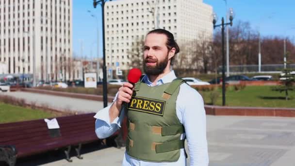 Medium shot, mannelijke televisiejournalist in een Bulletproof jasje praten met een microfoon in de voorkant van wolkenkrabbers in het financiële district van de stad. — Stockvideo