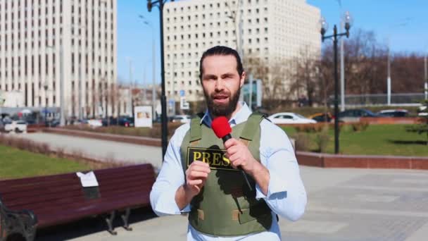 Medium shot, mannelijke televisiejournalist in een Bulletproof jasje praten met een microfoon in de voorkant van wolkenkrabbers in het financiële district van de stad. — Stockvideo