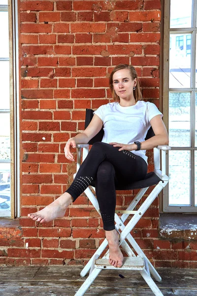 Hermosa jovencita descarada está sentada en una silla junto a la pared de ladrillo. ladrillo de fondo, espacio de copia — Foto de Stock