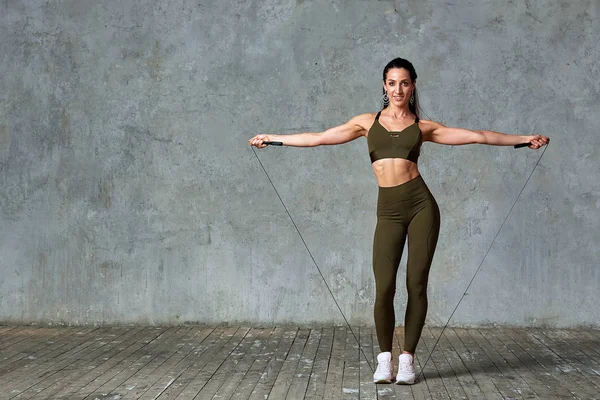 Modelo de aptidão sorridente posando no ginásio de comprimento completo contra uma parede cinza com jumpers em suas mãos, conceito Fitness — Fotografia de Stock