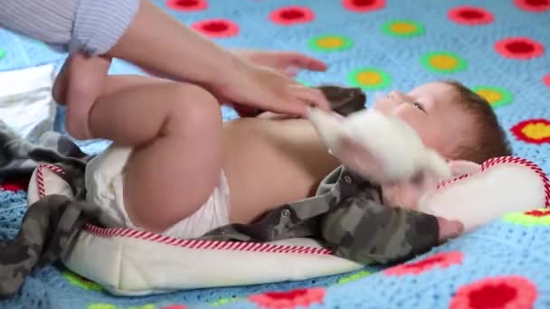 Moeder verwijdert kleren van een baby. Baby speelt met een zacht speelgoed terwijl liggend op een bed. Kleren worden van hem verwijderd. — Stockvideo