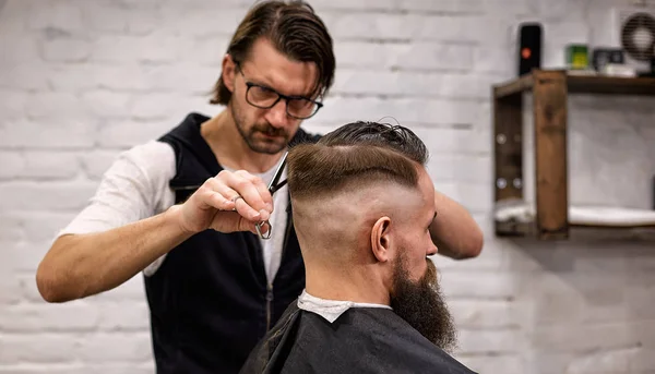 Master cuts hair and beard of men in the barbershop, hairdresser makes hairstyle for a young man