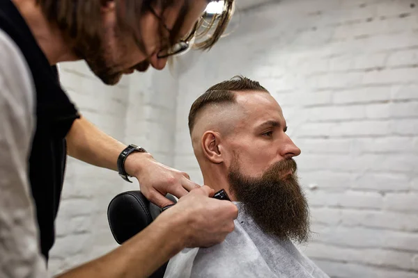 Мастер стрижет волосы и бороду мужчин в парикмахерской, парикмахер делает прическу для молодого человека — стоковое фото