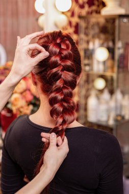Beautiful, red-haired girl with long hair, hairdresser weaves a French braid, in a beauty salon. Professional hair care and creating hairstyles. clipart