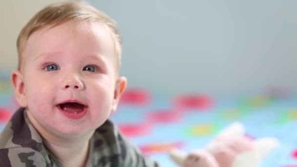 Hermoso bebé sonriente: Un bebé precioso se encuentra en la cama y sonríe a la cámara con un fondo de enfoque suave agradable — Vídeos de Stock