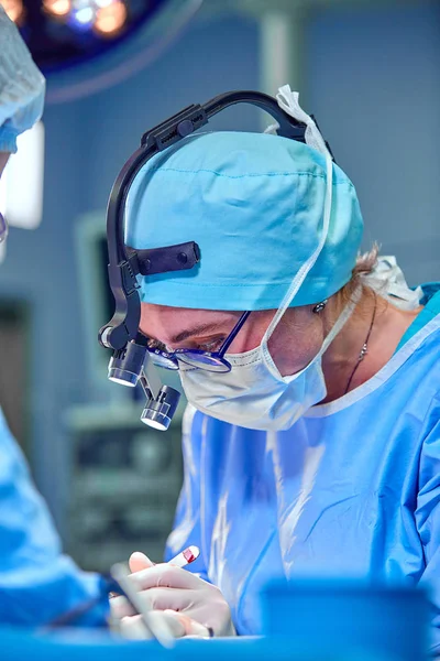 Surgeon and his assistant performing cosmetic surgery in hospital operating room. Surgeon in mask wearing loupes during medical procadure. — Stock Photo, Image